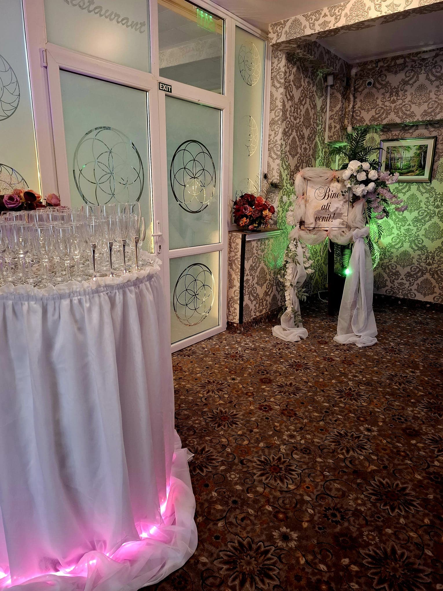 Intrare de eveniment cu mesajul Bine ați venit, flori albe și masă cu pahare de șampanie